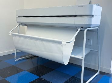 Lionel A large format printer on a blue and black checkered floor, positioned against a white wall, with rolled paper underneath.