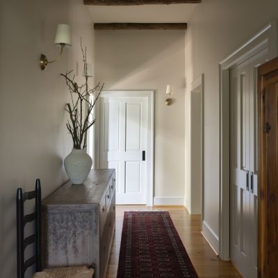 A rustic hallway features wooden ceiling beams, a red patterned rug, a vase with branches, a chair, and a pair of cowboy boots.