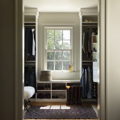 A neatly organized walk-in closet with hanging clothes, a window with natural light, and a colorful patterned rug on the wooden floor.