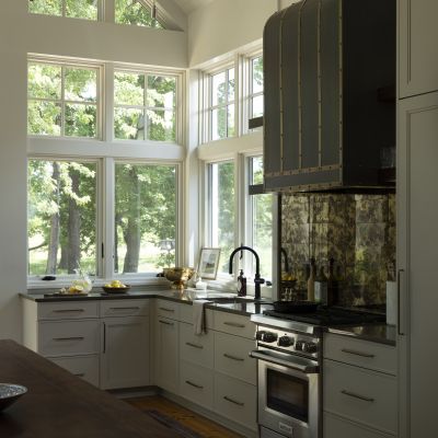 A modern kitchen with large windows, showcasing a sleek stove, dark countertops, reflective backsplash, and ample natural light through the trees outside.