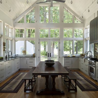 Spacious kitchen with high ceilings, large windows, wooden floors, long table, benches, and modern appliances. Abundant natural light illuminates the room.