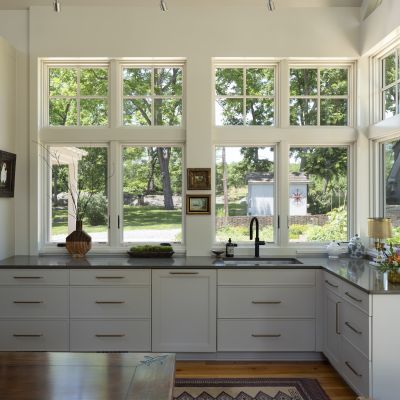 Sunlit kitchen with large windows, white cabinets, and black faucet. View of garden outside. Wooden table and decorative items inside.