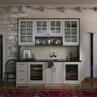 A cozy kitchen with rustic stone walls, featuring white cabinets, a wine cooler, and a variety of bottles on display.
