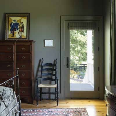 A cozy room features a wooden dresser, artwork, a black chair, colorful rug, and glass door overlooking greenery outside.