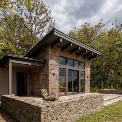Modern, single-story house with stone exterior, large windows, and a patio. Surrounded by lush trees under a partly cloudy sky.