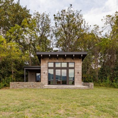 Modern small house with large windows in a grassy field, surrounded by lush trees under a partly cloudy sky.