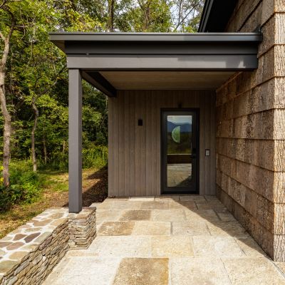 Modern entrance with stone pathway and rustic wood siding, surrounded by trees. Glass door reflects outdoor scenery, creating a peaceful, natural ambiance.