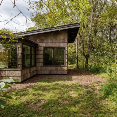 A modern house with large windows is nestled among lush trees, blending harmoniously with the natural forest surroundings on a sunny day.