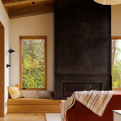Cozy living room with a modern fireplace, wooden accents, large window views, and a red sofa draped with a patterned blanket.