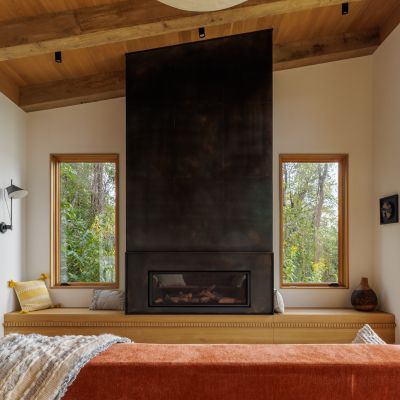 Modern living room with a large central fireplace, wooden ceiling beams, and views of lush greenery through tall windows. Cozy seating with cushions.