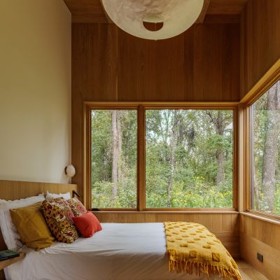 A cozy wooden bedroom with large windows reveals a lush green forest outside. The bed features colorful pillows and a yellow throw.