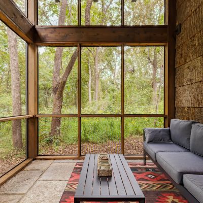 A cozy screened porch with large windows, a couch, and a patterned rug, overlooking a serene, wooded landscape.