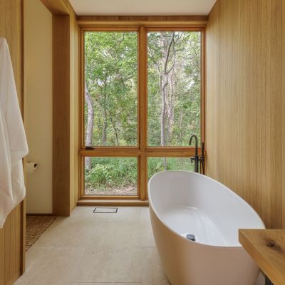 A serene bathroom with a standalone white tub, wooden walls, and large windows offering a view of lush greenery outside.
