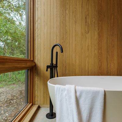 A minimalist bathroom features a wooden wall, freestanding bathtub with a towel draped over, and a window overlooking a natural, leafy landscape.