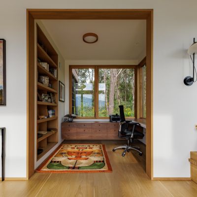 A modern home office features large windows, a vibrant rug, built-in shelves, and scenic nature views, creating a serene, organized workspace.