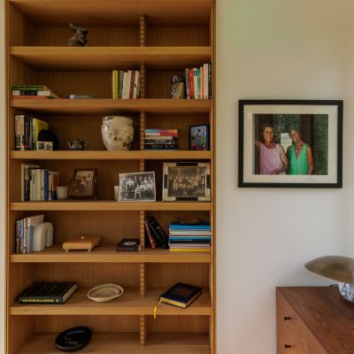 A neatly arranged wooden bookshelf with books, framed photos, and decorative items is next to a photograph of two people on the wall.