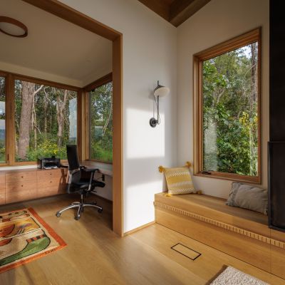 Modern room with wooden floors, desk, and chairs. Large windows reveal a forest view, while a colorful rug adds warmth. Cozy ambiance.