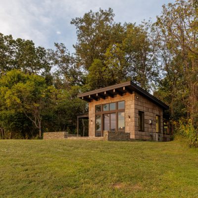 A modern cabin with large windows is nestled among lush trees on a grassy hillside, bathed in soft afternoon light.