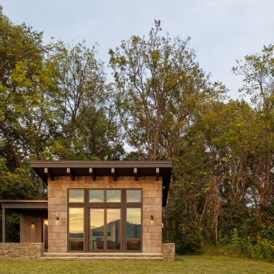 A modern, single-story house with large windows sits in a grassy clearing, surrounded by tall trees under a clear sky.