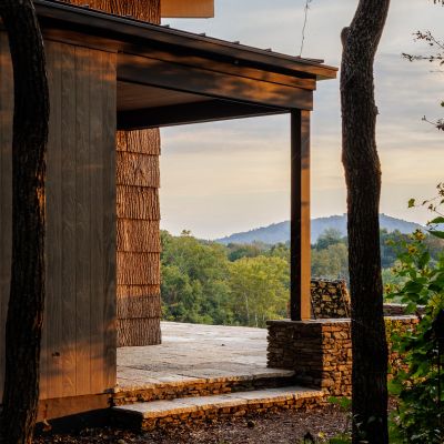A modern building with wooden and stone elements in a serene, wooded landscape with hills in the background during sunset.
