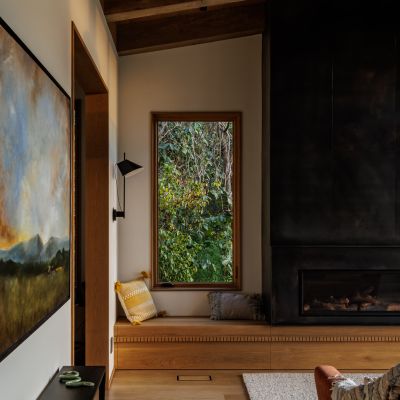 Cozy modern room with wooden elements, a window seat, and a fireplace. Large landscape painting and soft lighting add warmth.