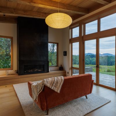 A cozy living room features large windows, a fireplace, and a view of lush greenery and distant mountains. Warm lighting and comfortable seating.