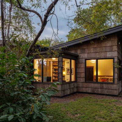 A cozy wooden house in a lush forest, with illuminated windows revealing a warmly lit interior during dusk.