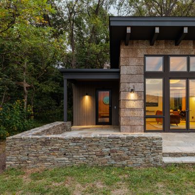 Modern house with large windows nestled in a wooded area. Exterior features stone accents and soft outdoor lighting. Elegant, natural setting.