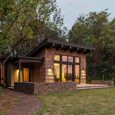 Modern cabin with large windows and illuminated interior, surrounded by lush greenery. Stone and wood exterior creates a warm, inviting atmosphere.