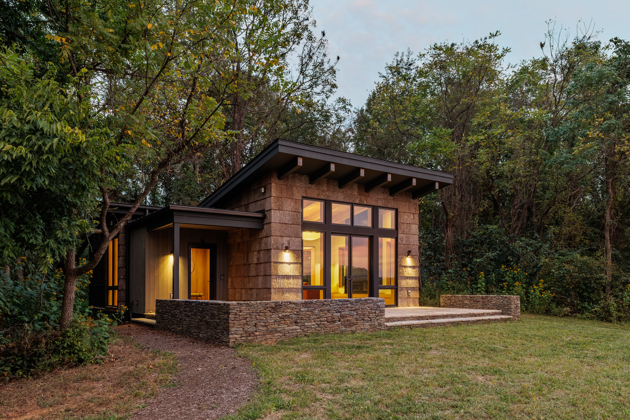 Modern cabin with large windows and illuminated interior, surrounded by lush greenery. Stone and wood exterior creates a warm, inviting atmosphere.