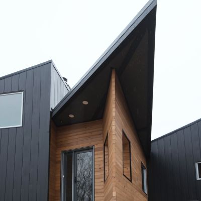 Modern house with sharp angles and contrasting black and wooden siding. Large windows illuminate the minimalist, geometric architecture, creating an inviting, sleek appearance.