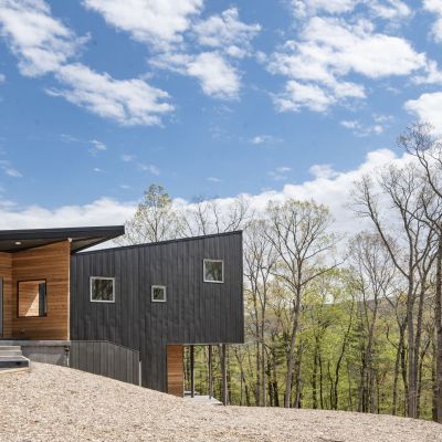 Modern, angular house with dark exterior, large windows, and wood accents amid forest setting. Clear blue sky with thin clouds above.
