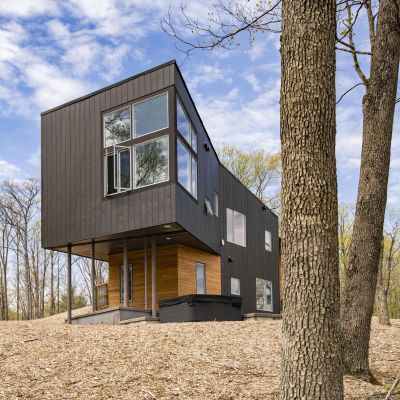 A modern, two-story black and wood house in a wooded area, surrounded by tall trees under a partly cloudy sky.