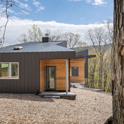 Modern cabin with metal and wood exterior nestled in a serene, wooded area. Trees surround the structure under a partly cloudy sky.