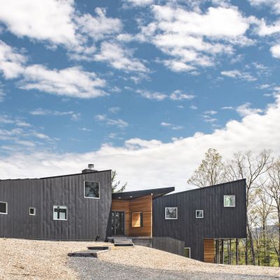 Modern, angular black house with large windows on a hilltop, surrounded by trees under a partly cloudy sky, showcasing minimalist architecture.
