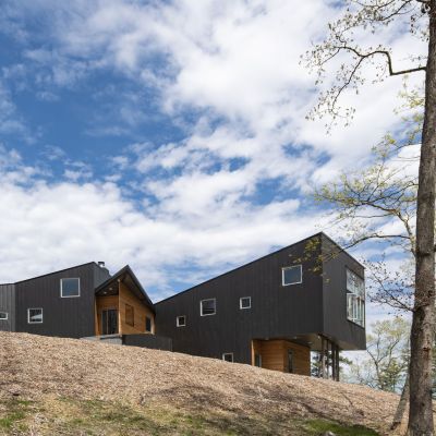 Modern black house on a wooded hill under a partly cloudy sky, surrounded by trees and natural scenery.