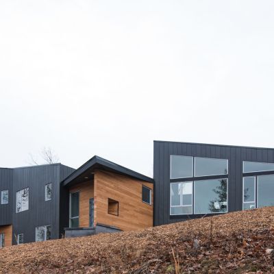 Modern, angular house on a sloped, leaf-covered terrain. Features dark panels and large windows. Surrounded by bare winter trees under a cloudy sky.
