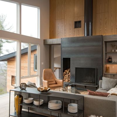 Modern living room with large windows, wooden ceiling, and a fireplace. A comfortable leather chair and tableware on open shelving complete the cozy setting.