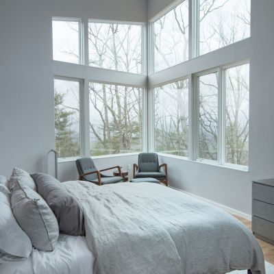 Modern bedroom with large windows, grey bedding, and wooden furniture. Chairs face a wintery tree view outside. Book on nightstand. Serene atmosphere.