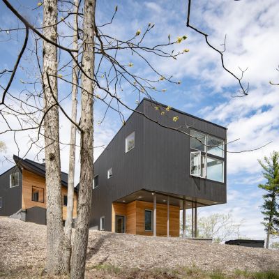 Modern, elevated house with black and wooden exterior surrounded by bare trees under a partly cloudy sky. No people or landmarks visible.