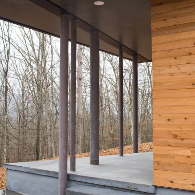 A modern porch with wooden and metal elements, surrounded by bare trees, features concrete steps and a sleek overhang in a forest setting.